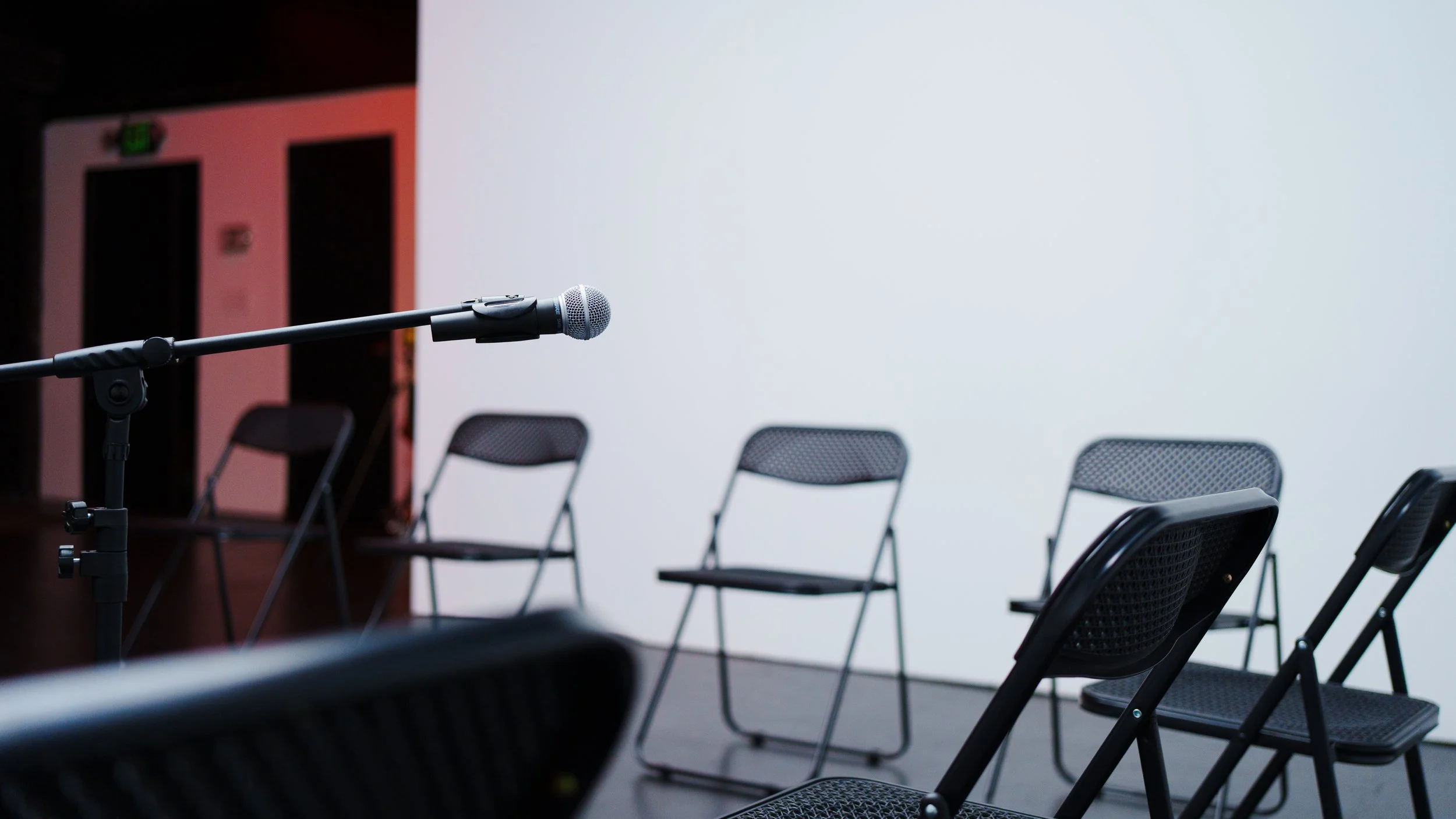 Empty conference room with black mesh chairs arranged in rows, a portable microphone on a stand, a white projection screen, and a pinkish wall in the background.