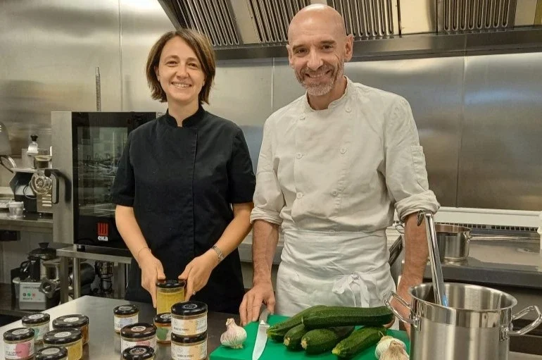 A woman in a black chef coat and a man in a white chef coat are standing in a commercial kitchen, smiling at the camera. On the counter in front of them are jars of sauces or condiments, zucchinis, garlic, and a container of water.