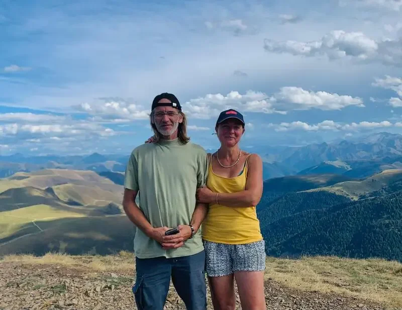 A man and woman standing together on a mountain with scenic rolling hills and distant mountains in the background under a partly cloudy sky.