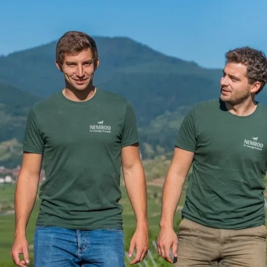 Two men walking outdoors on a sunny day, wearing green t-shirts with a logo and text, with mountains and fields in the background.