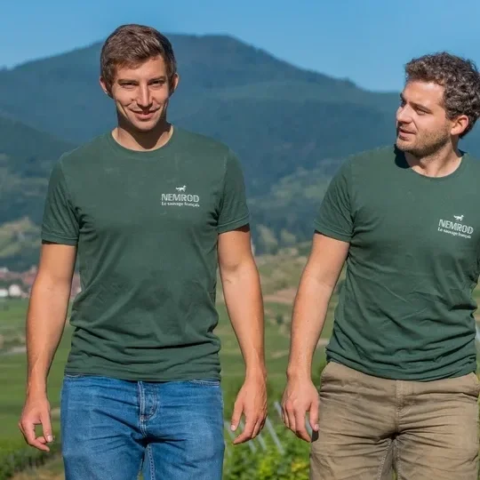 Two men walking outdoors in a rural area with hills in the background, both wearing green T-shirts with white text and logo.