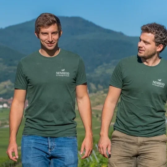 Two men in matching green T-shirts walking outdoors with mountains and green fields in the background.