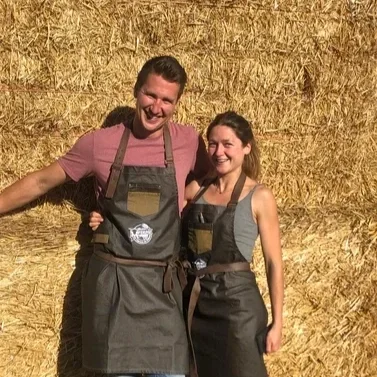 Two smiling people, a man and a woman, standing in front of a haystack, wearing aprons, on a farm or in a rural setting.