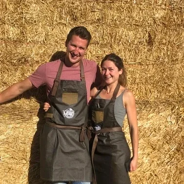 A smiling man and woman stand close together in front of a straw wall, both wearing black aprons with pockets and straps, appearing to be on a farm or outdoor setting.