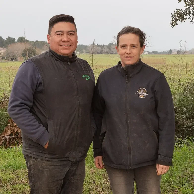 A man and woman standing outdoors in a grassy field with trees and fields in the background, wearing black jackets.