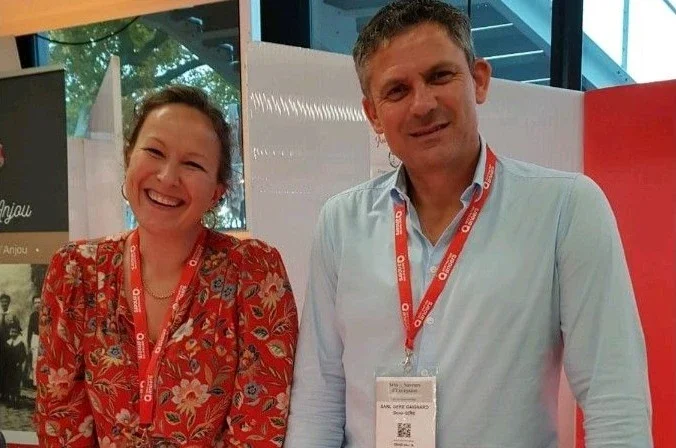 A smiling woman with brown hair and a man with short dark hair, both wearing conference badges and lanyards, standing indoors in front of a red and white backdrop.