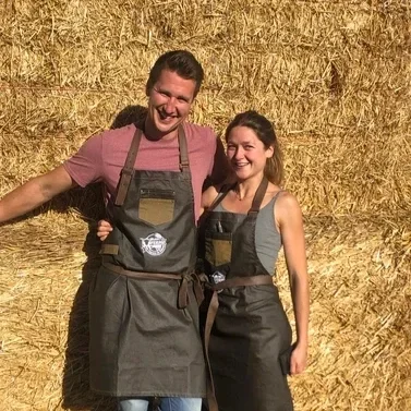 Two people, a man and a woman, smiling and posing in front of a large haystack, wearing aprons.