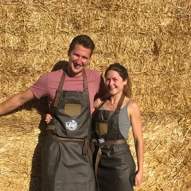 Two smiling people, a man and a woman, wearing apron and standing in front of a haystack.