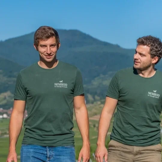 Two men wearing green Nemrod T-shirts walking outdoors with mountains and greenery in the background.
