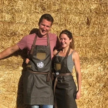 A smiling man and woman wearing aprons, standing in front of a haystack outdoors.