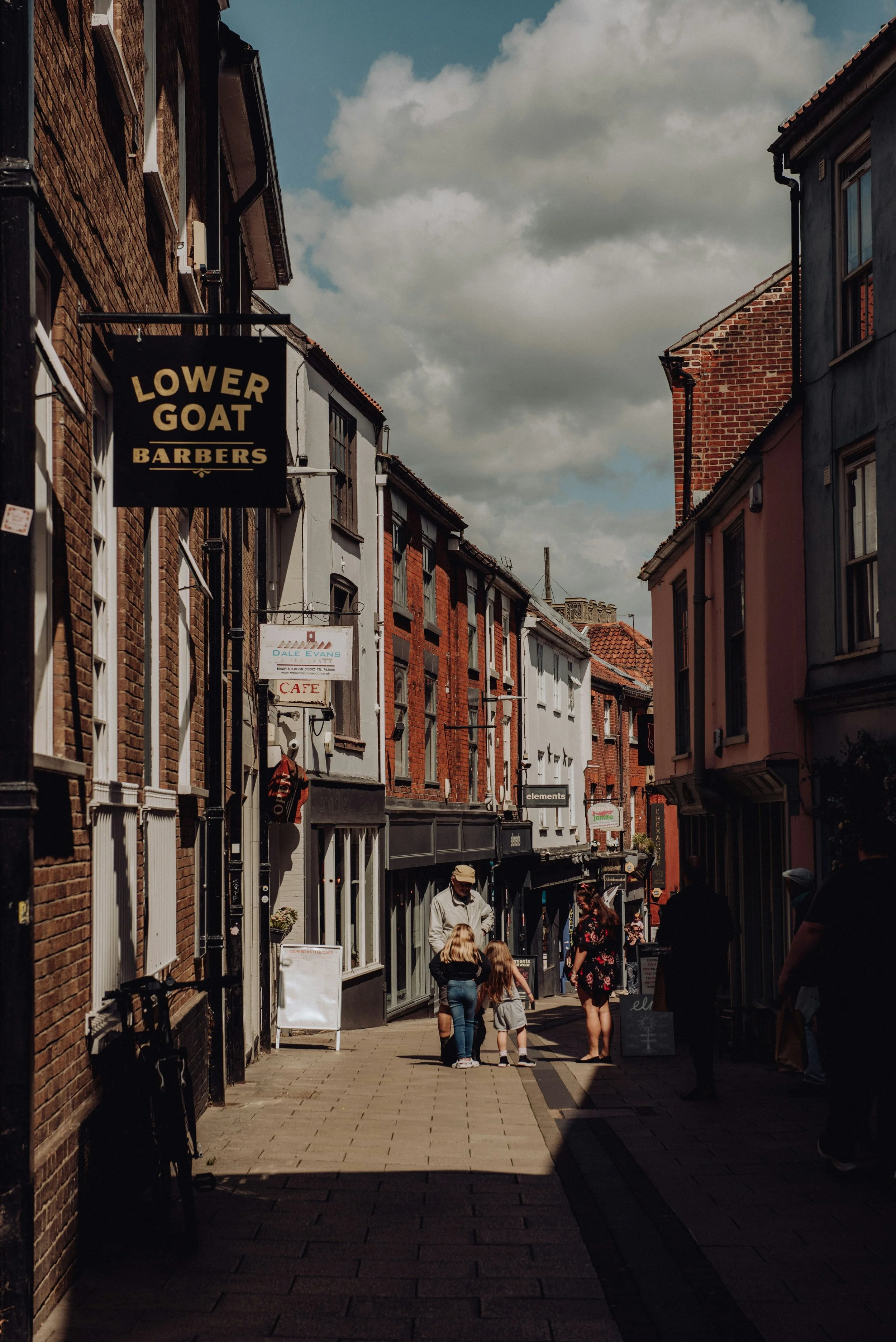 A street scene with people walking along a narrow sidewalk, lined with brick and painted buildings under a partly cloudy sky. Signs for a barbershop and a cafe are visible.