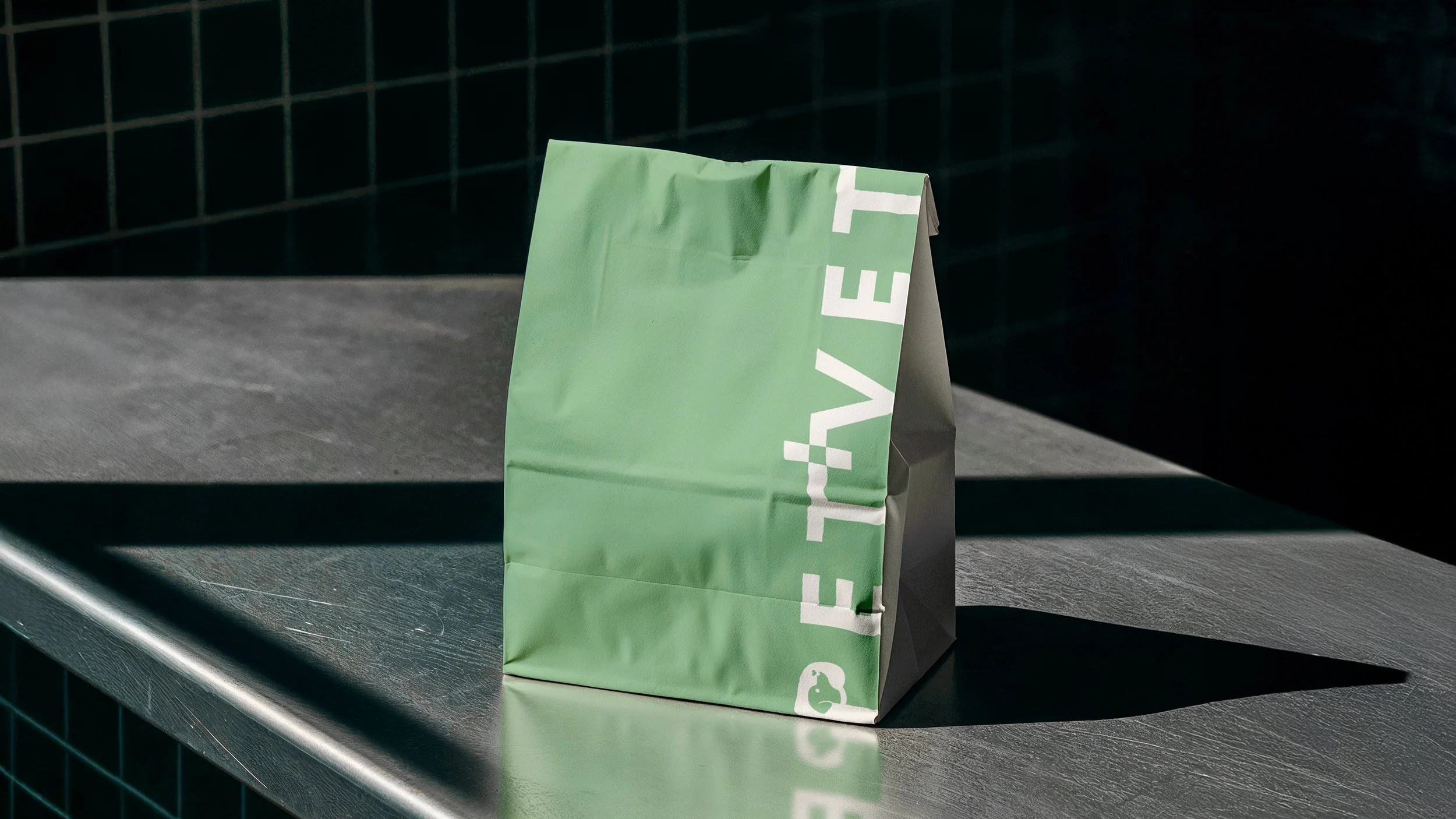 A green paper bag with white text reading 'EAT' standing on a dark metallic surface, casting a shadow to its right, with a dark tiled wall in the background.