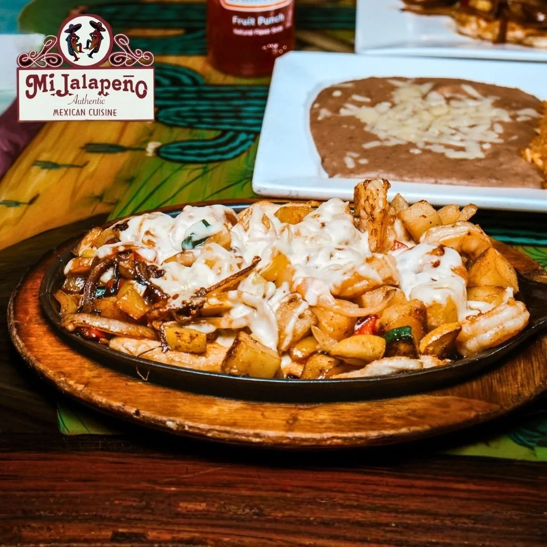 Mexican dish with mixed grilled vegetables topped with melted cheese, served on a black plate with a wooden base. In the background, there is a split serve of refried beans topped with cheese and a sign for Mi Jalapeno Mexican Cuisine.