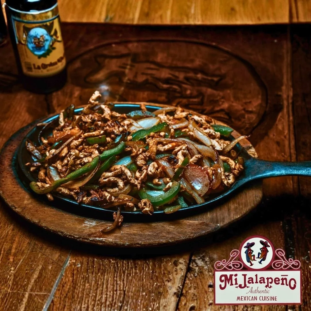 A sizzling plate of beef stir-fry with green bell peppers and onions, served on a wooden platter, with a bottle of hot sauce labeled 'La Chupacabra' in the background. The image also features a logo for Mi Jalapeño Mexican Cuisine.