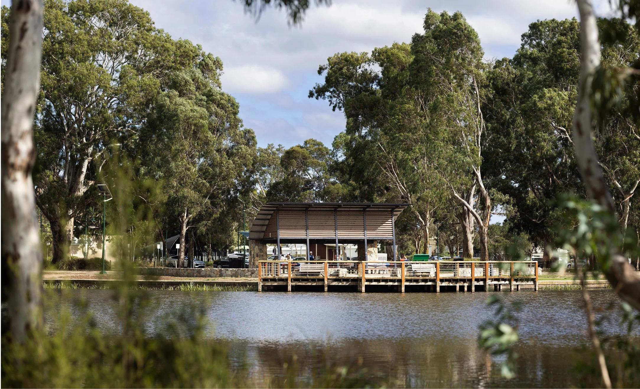 A park with a wooden shelter on a pier over Banjo Patterson lake, surrounded by tall green trees and a cloudy sky.