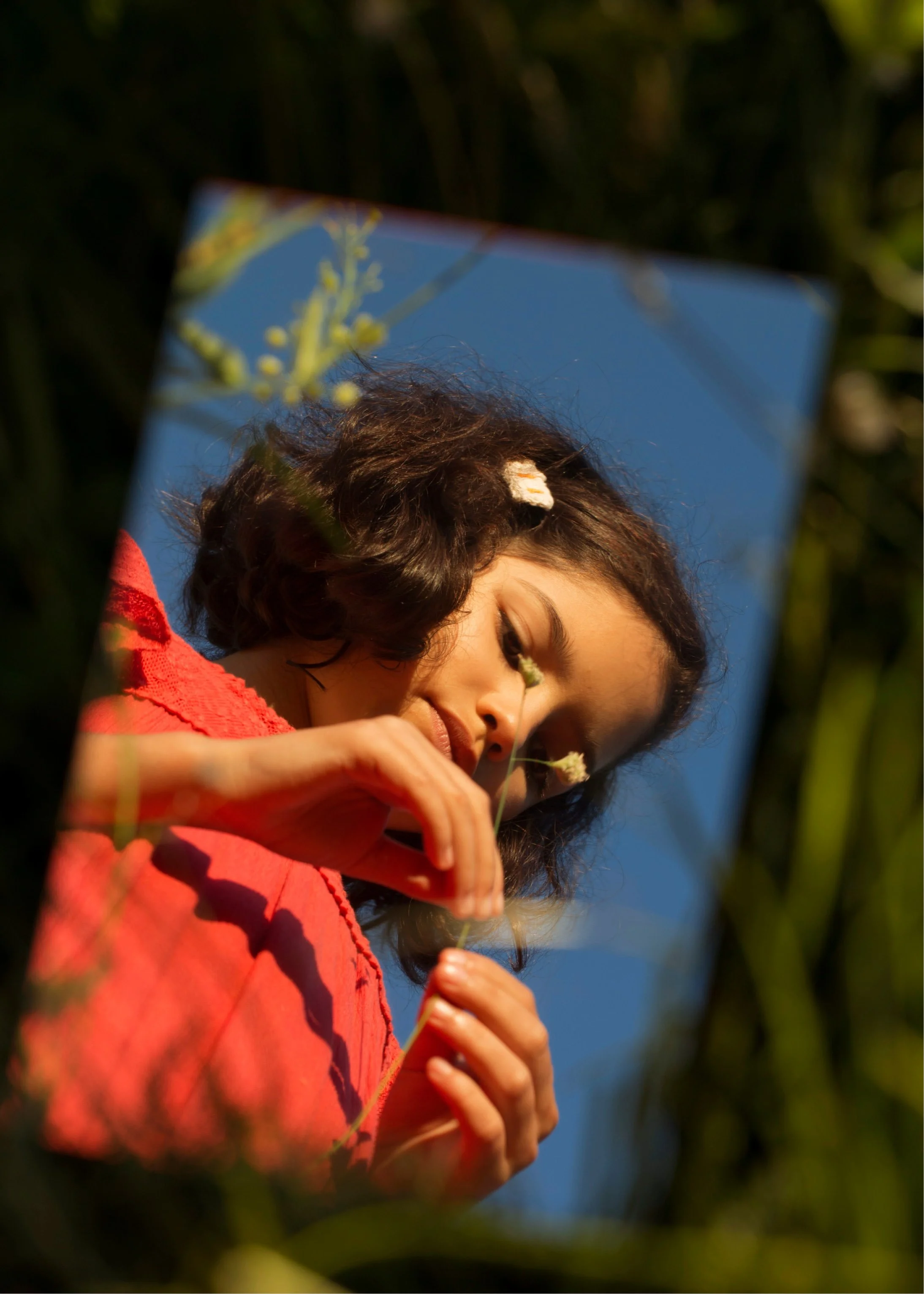 A young woman with dark, curly hair and light skin is outdoors, inspecting or smelling a small flower she is holding, with a clear blue sky in the background.