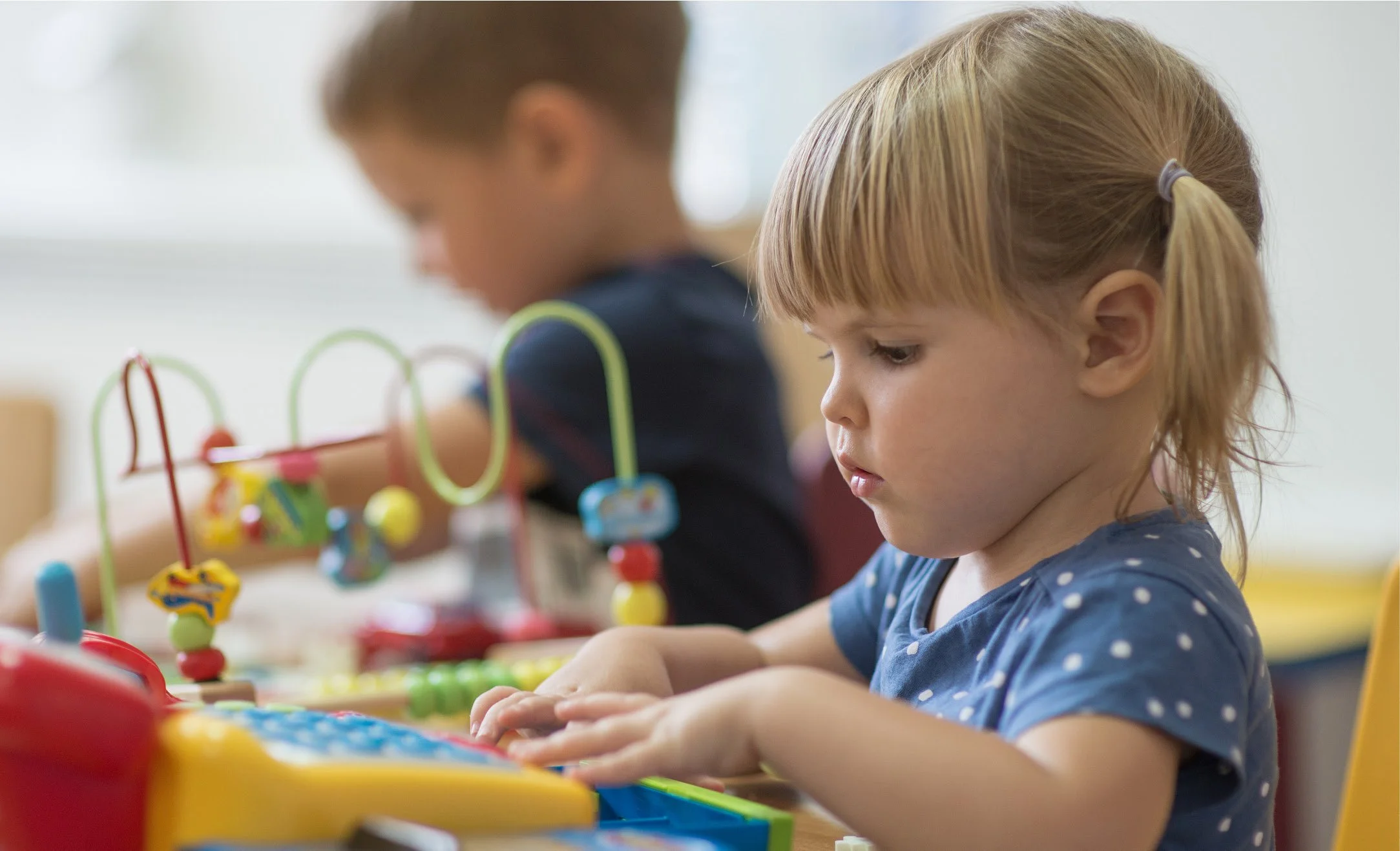 Two young children, a girl with red hair and a boy with light brown hair, are focusing on playing with a colorful educational toy on a table. The girl wears a blue polka dot dress, and the boy wears a dark shirt.