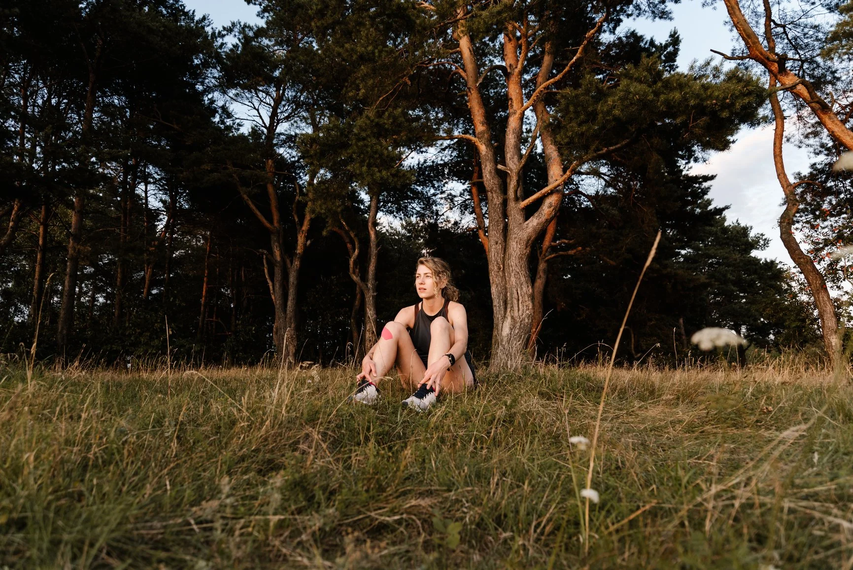 A young woman in athletic clothing sitting on the grass in a park, tying her shoelaces, with tall trees in the background at dusk.