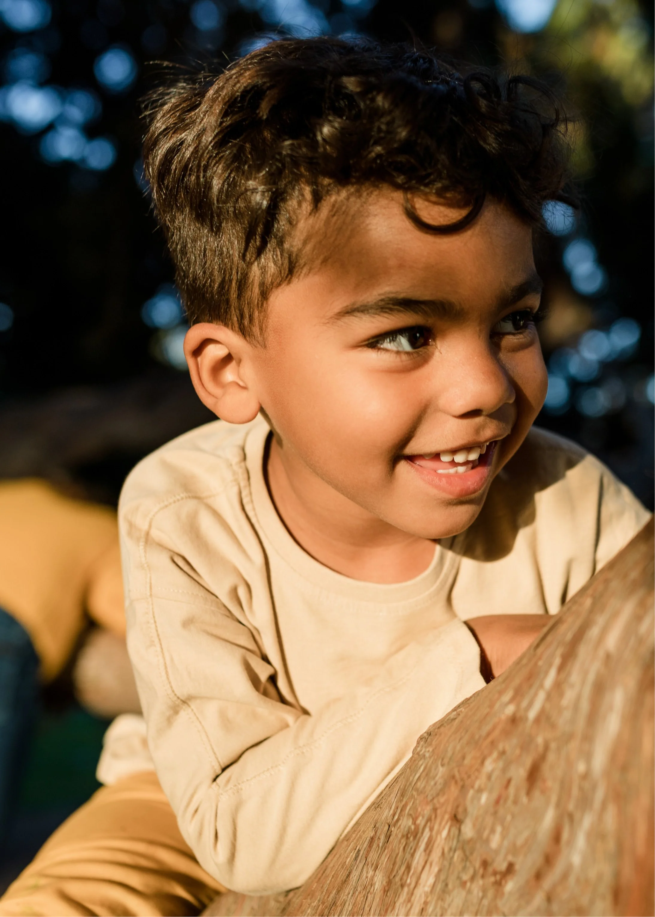 A young boy with curly dark hair smiling and playing outdoors on a tree branch during daytime.