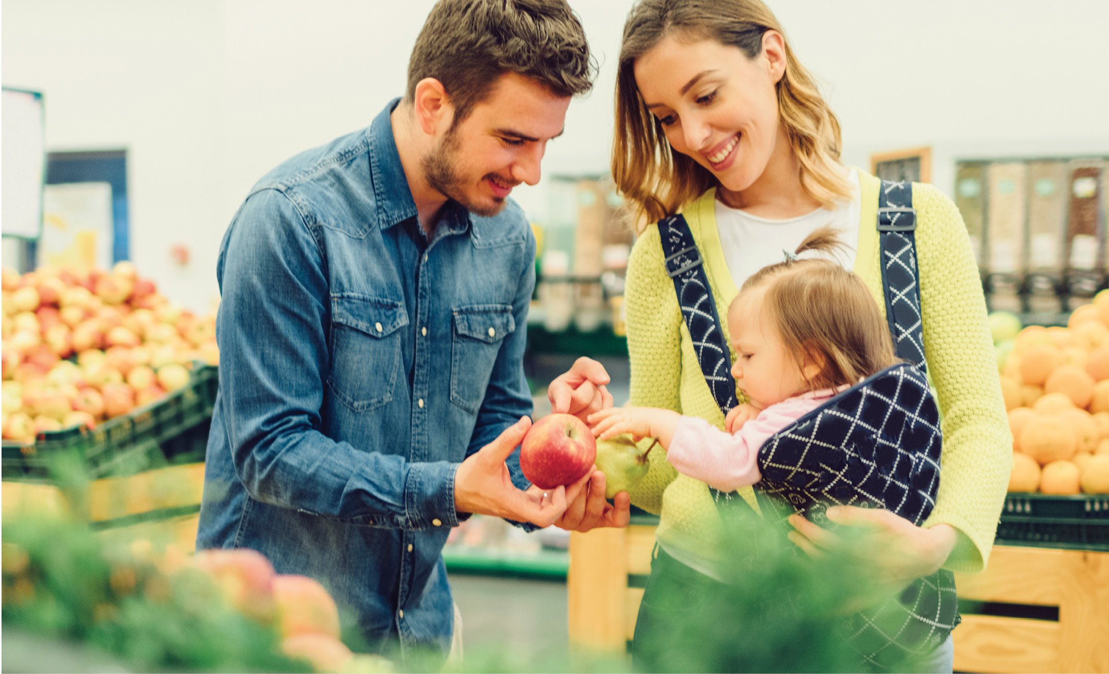A family shopping for apples at a grocery store, with a man holding an apple, a woman carrying a baby, and a toddler reaching toward the apples.