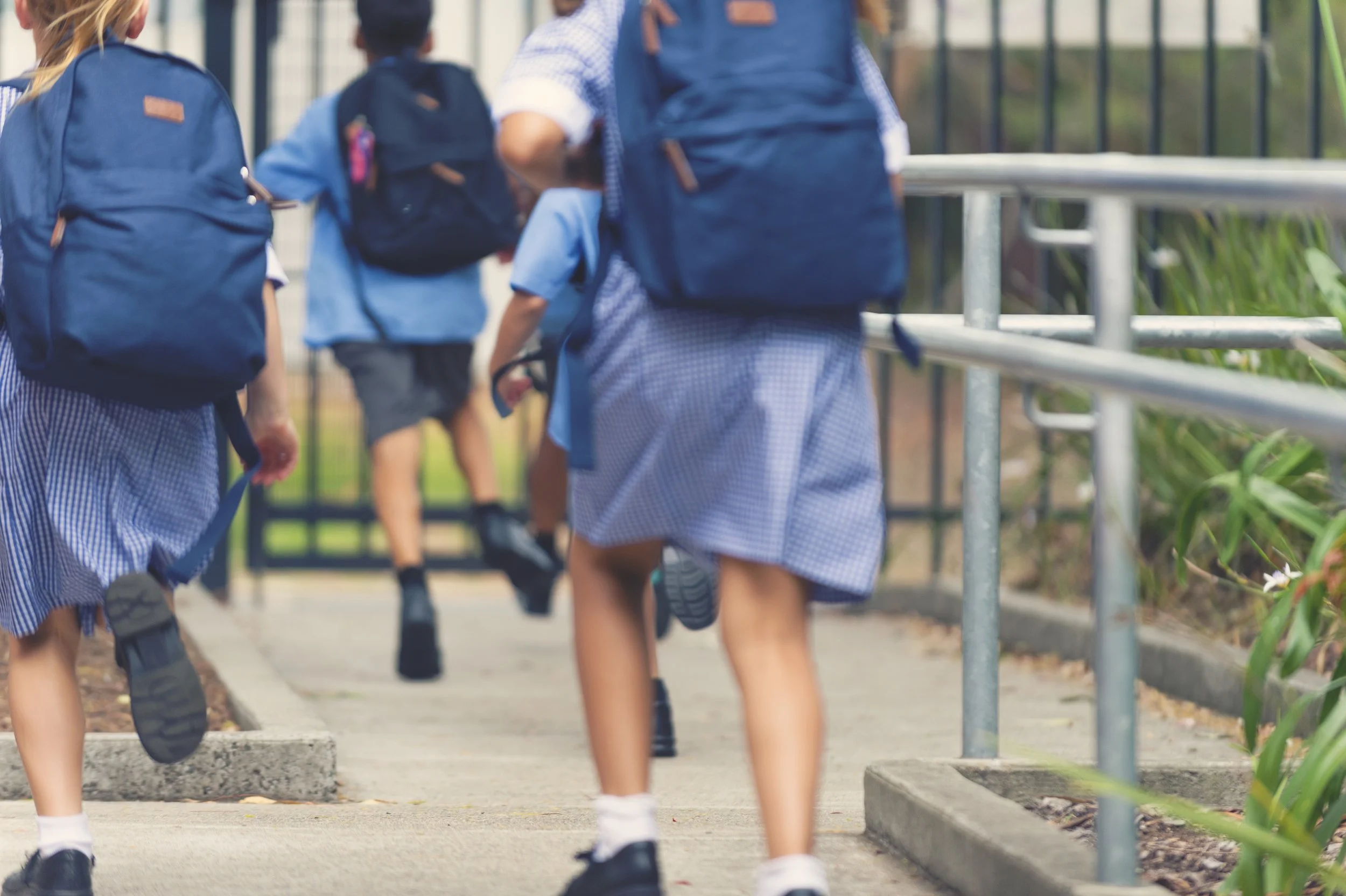 Children in school uniforms carrying backpacks, walking up stairs toward a gated entrance.