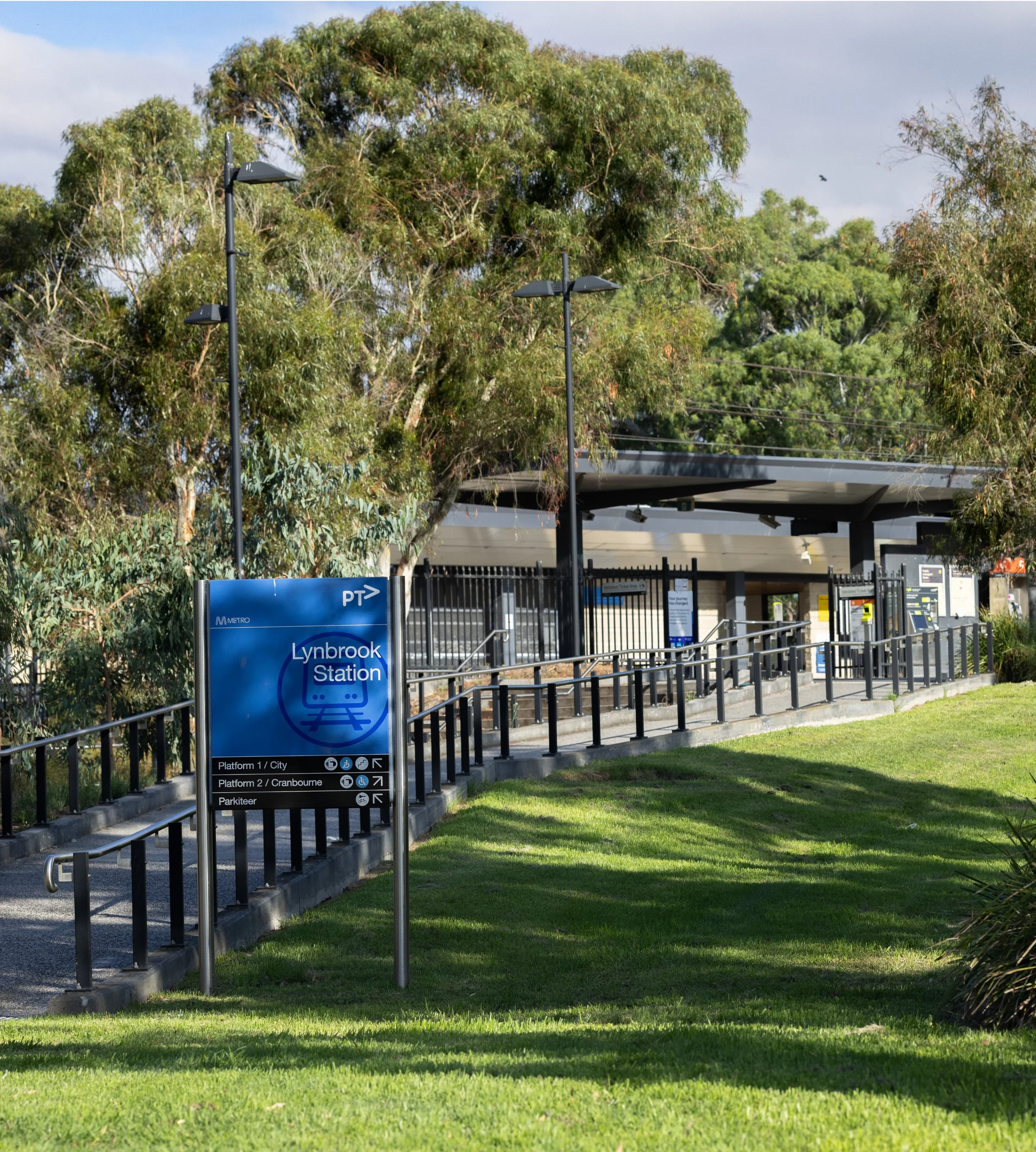 Lynbrook Train Station entrance with stairs and ramps, surrounded by green trees and grass, and a blue sign with station information.