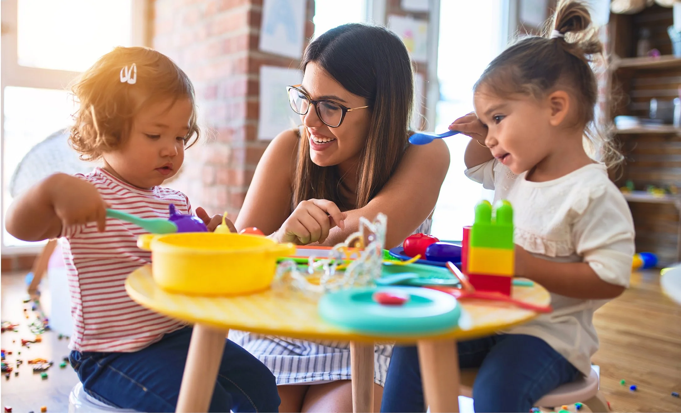 A woman with glasses and two young girls play with colorful Play-Doh at a small table in a bright room, with a brick wall and shelves in the background.