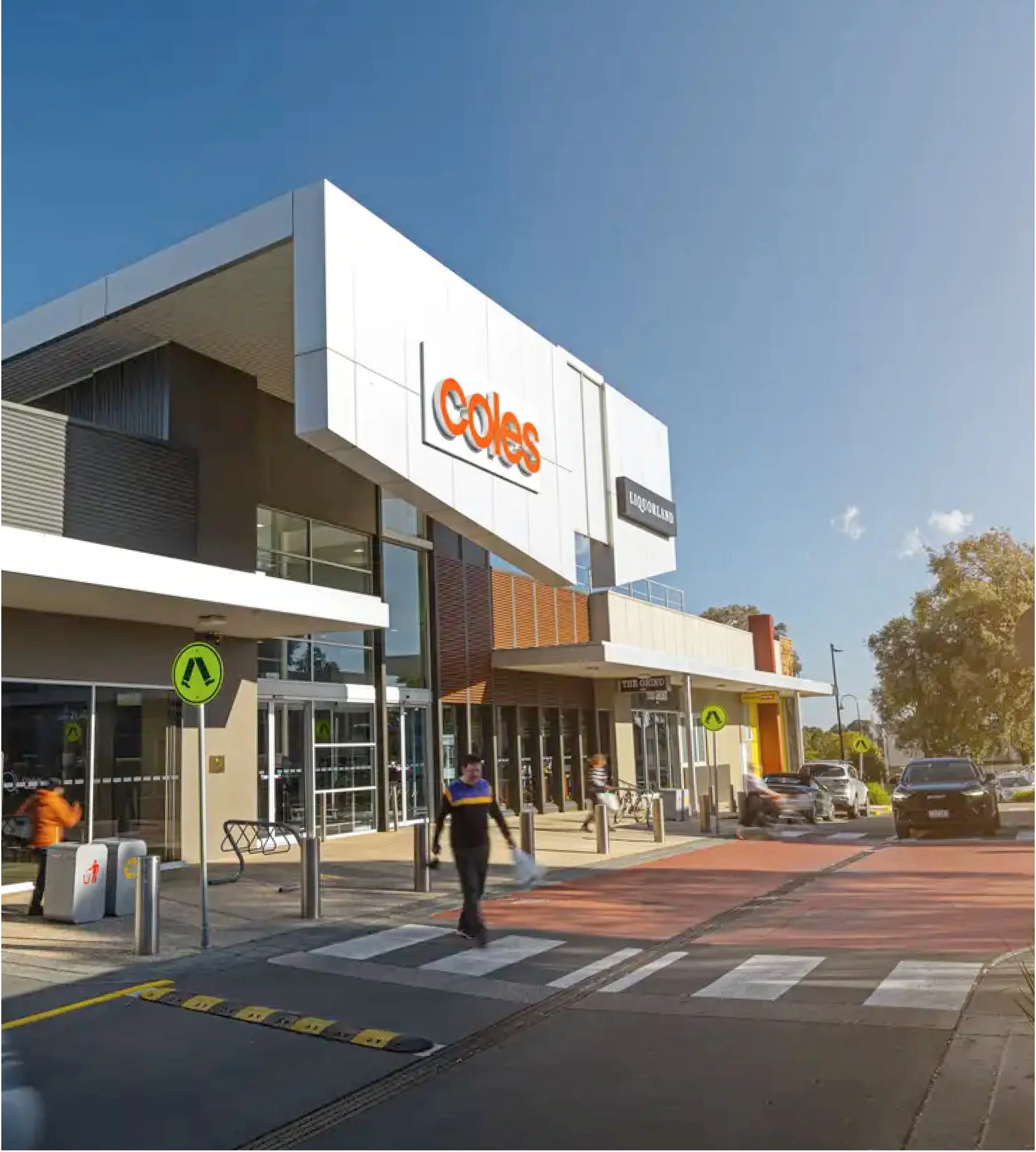 The image shows the exterior of Lynbrook Shopping Village, with a prominent sign for Coles. People are walking near the entrance, and cars are parked or driving in the parking lot. There are yellow pedestrian crossing signs and a crosswalk in front
