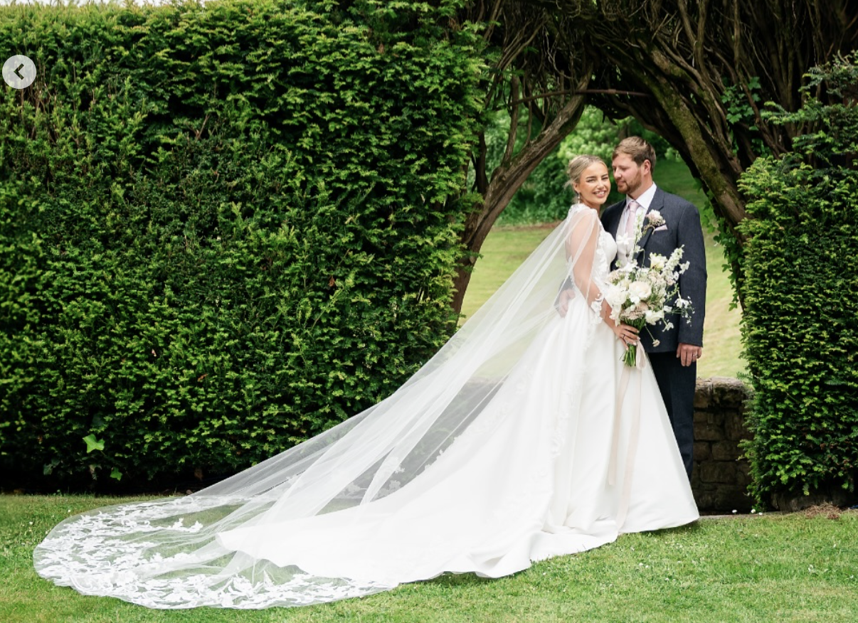 A bride and groom standing together outdoors under a natural archway of trees, dressed in wedding attire, smiling and holding a bouquet of flowers.
