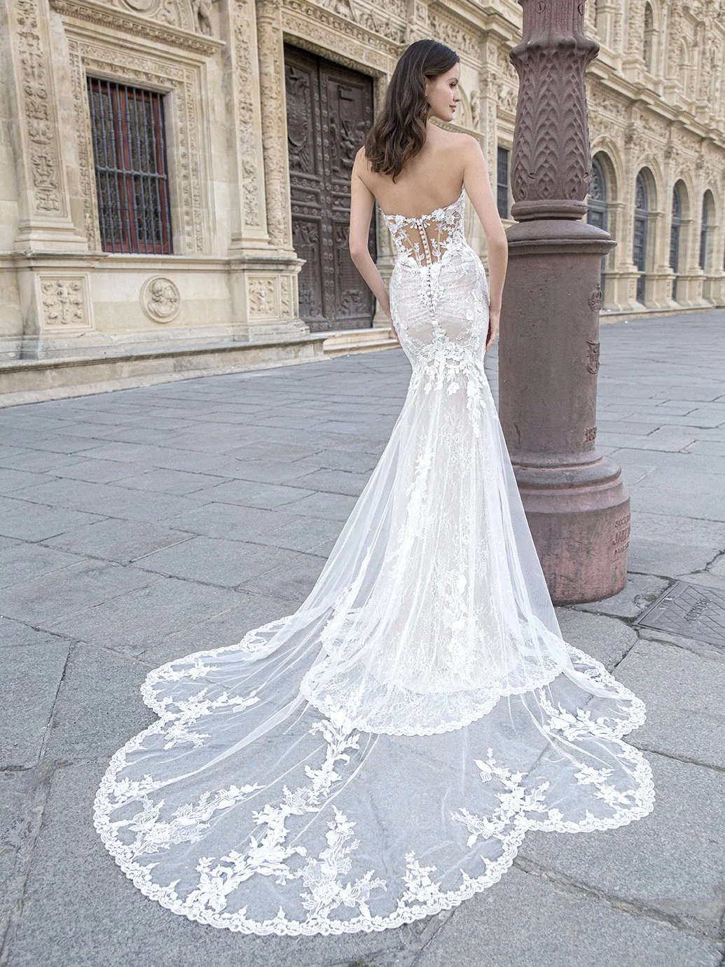 A woman in a white, lace wedding gown with a long train stands on a city sidewalk, facing away from the camera, near an ornate building and a decorative streetlamp.