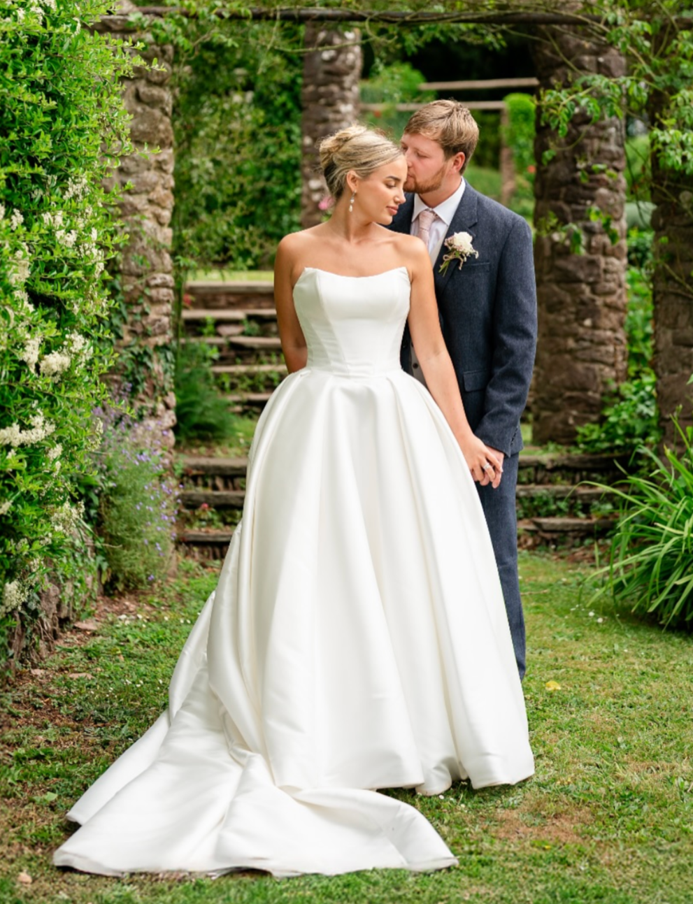 Bride in a white wedding gown and groom in a dark suit with a boutonniere, standing close in a garden with stone pillars, lush greenery, and wooden stairs in the background.