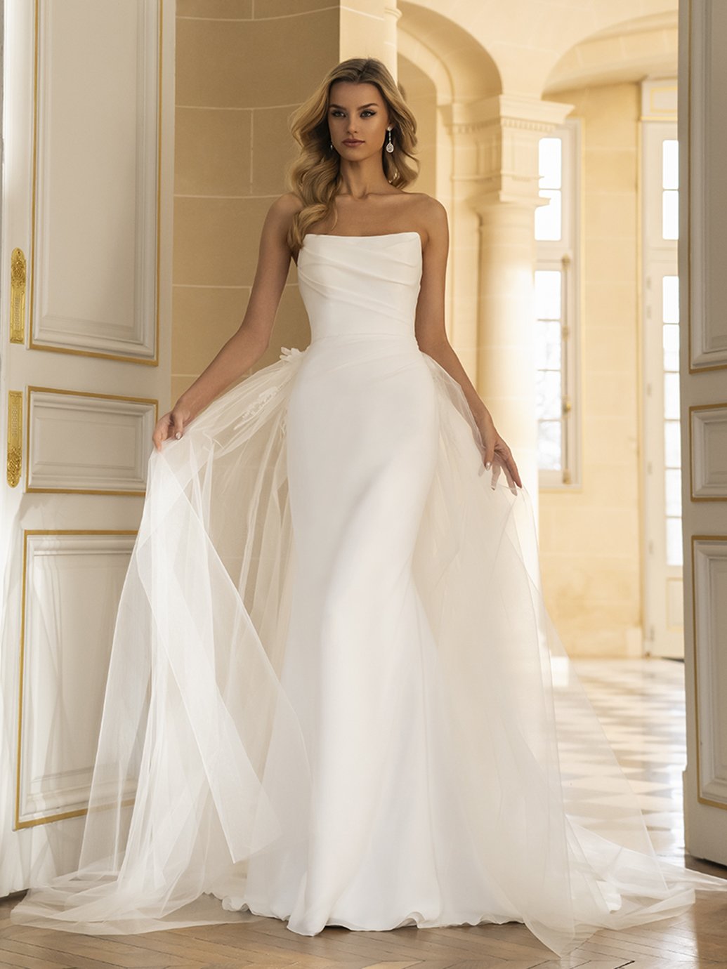 A woman in an elegant white strapless wedding gown with a full skirt and tulle overlay, standing in a grand, well-lit room with cream-colored walls and ornate molding.