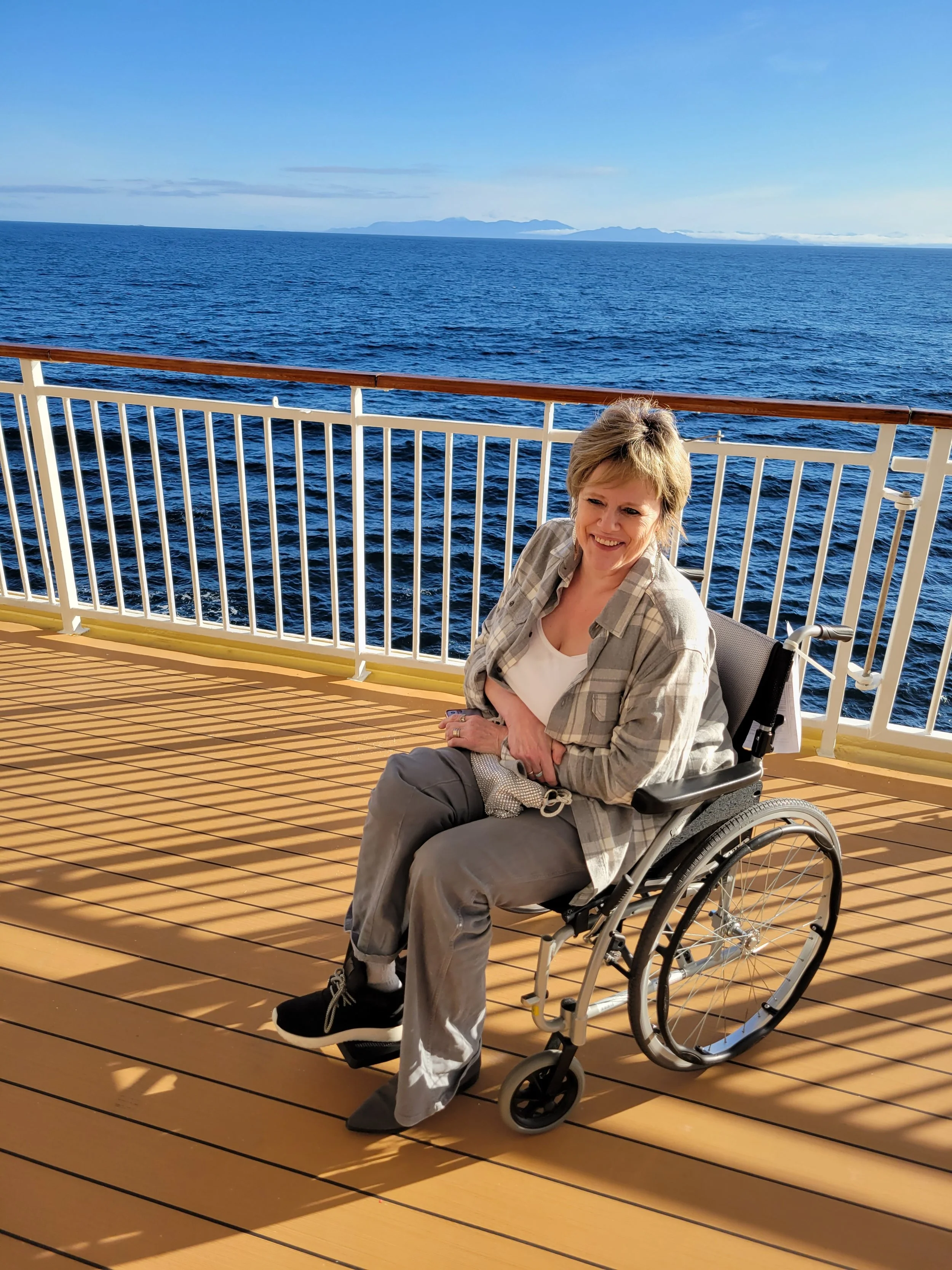 The sun is shining on Sandy in her wheelchair on the deck of a cruise ship in front of the blue ocean