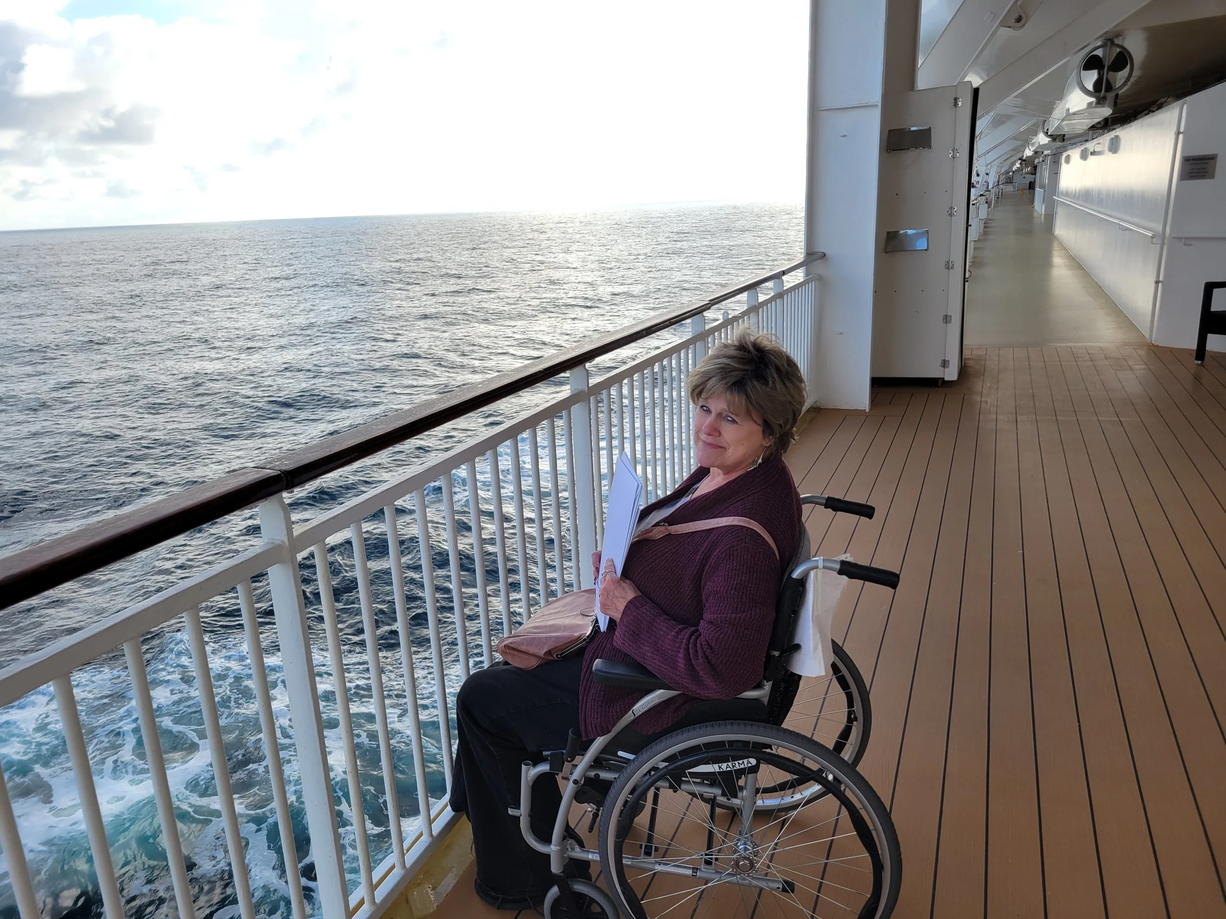 The sun is shining on Sandy in her wheelchair on the deck of a cruise ship in front of the blue ocean