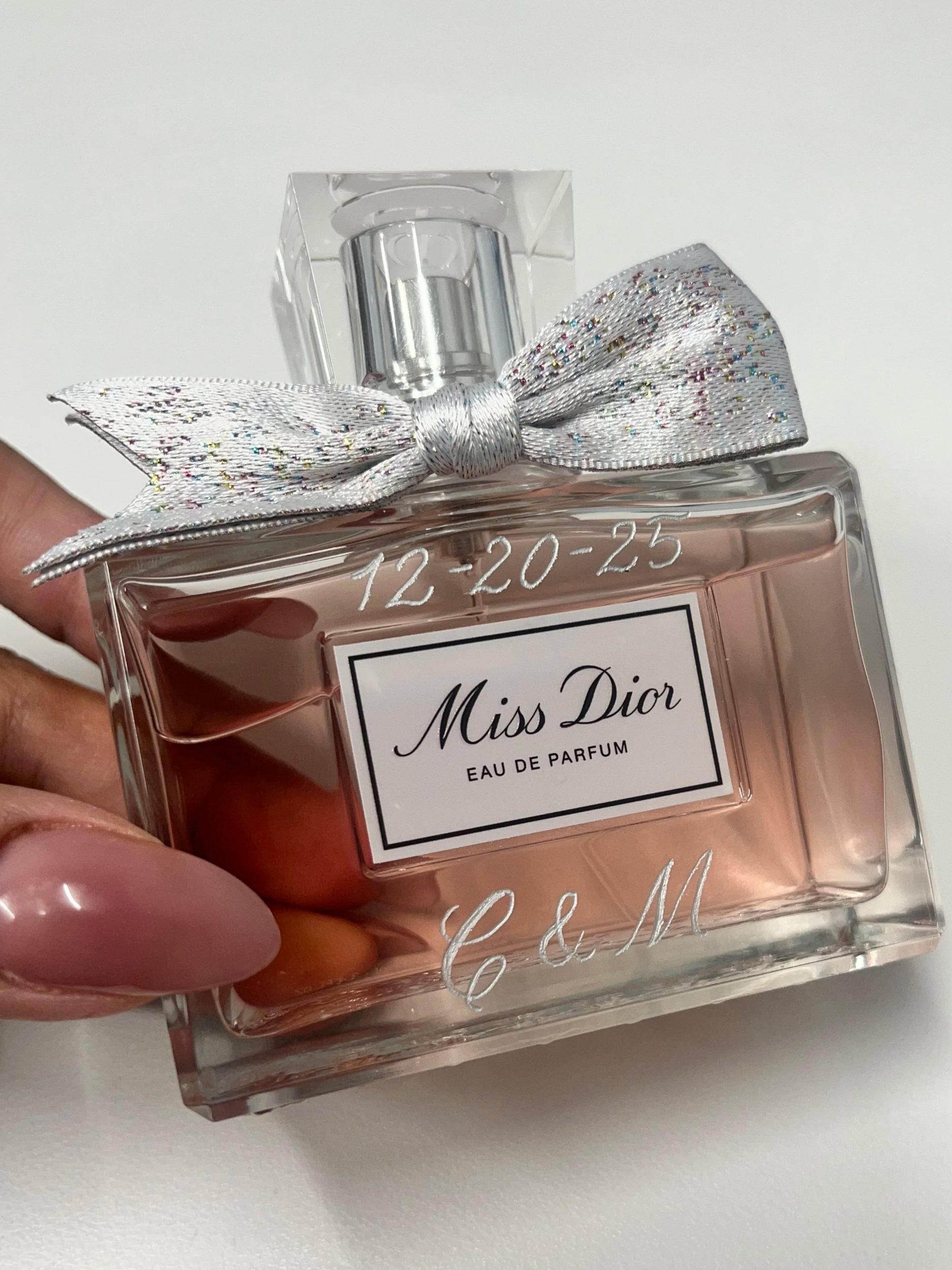 A person holding a bottle of Miss Dior Eau De Parfum perfume with a silver glittery bow on top and the date '12-20-25' custom enrgaved on the bottle.