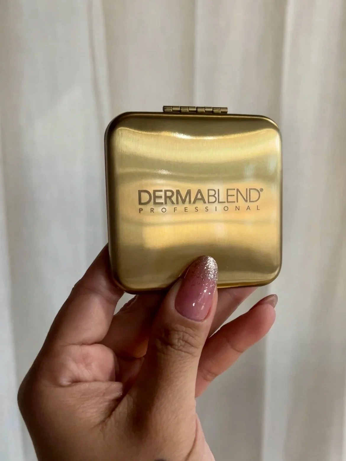 A hand holding a small, gold-colored compact case with 'DERMABLEND PROFESSIONAL' engraved on it.