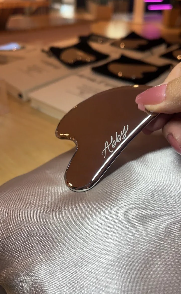 Close-up of a gold-colored nail file with the word 'Abyss' engraved on it, held by a person with pink nail polish. In the background, there are blurred items and a wooden surface.