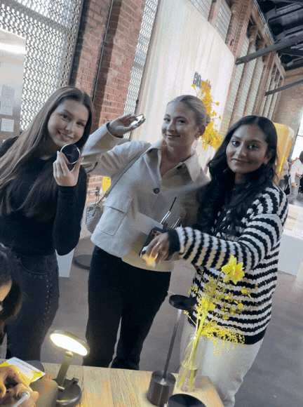 Three women smiling and posing in an industrial-style indoor space, with a table and yellow flowers in a vase in the foreground.