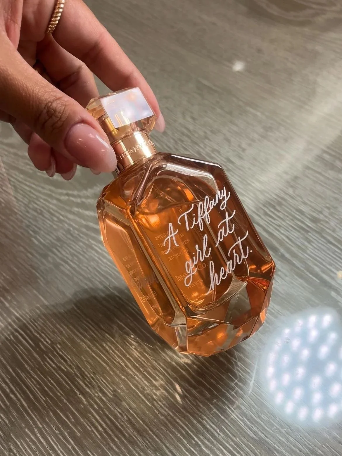 A hand holding a bottle of perfume with the phrase "A Tiffany girl at heart" written on it, placed on a wooden surface.