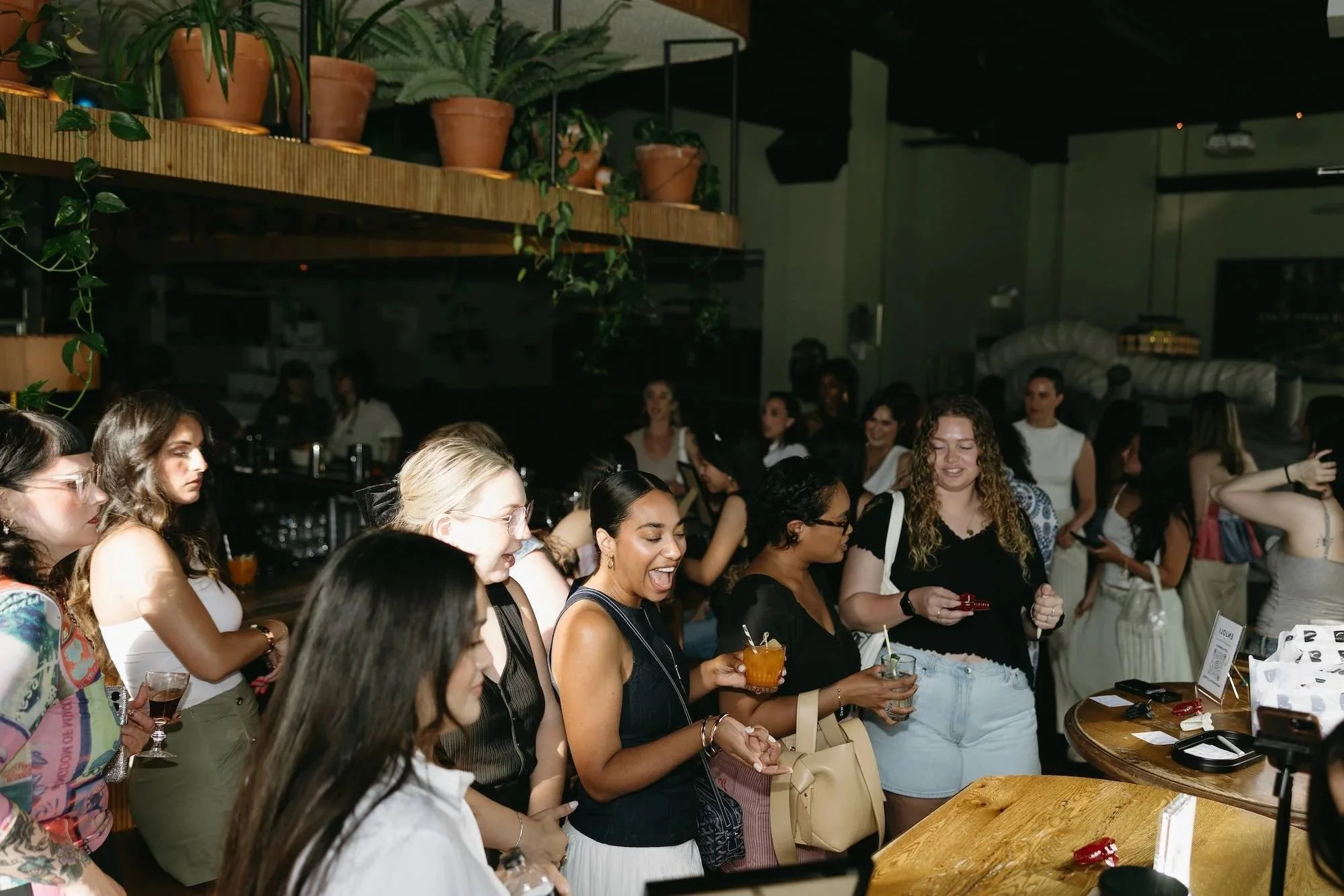 A group of women at a social gathering inside a dimly lit venue, some holding drinks, with plants and wooden shelves in the background.