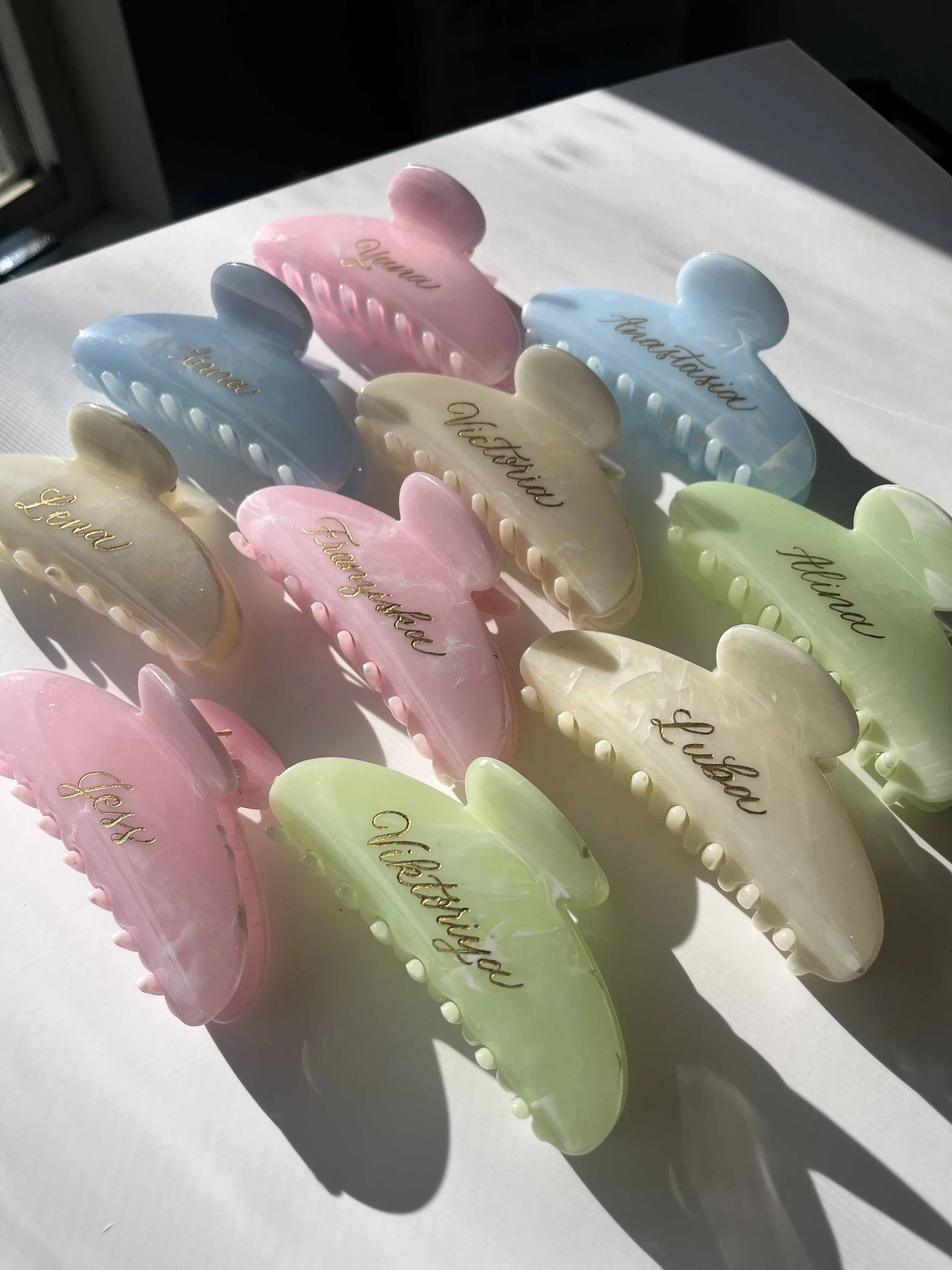 Colorful butterfly-shaped hair clips with personalized names written in gold script, arranged on a white surface in sunlight.