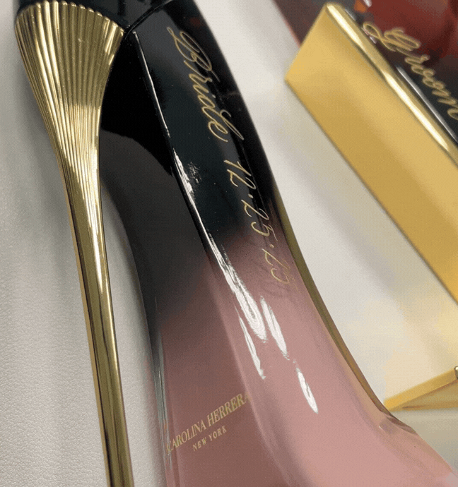 Close-up of a Carolina Herrera perfume box with a gold and black design, and a gold perfume bottle in the background.