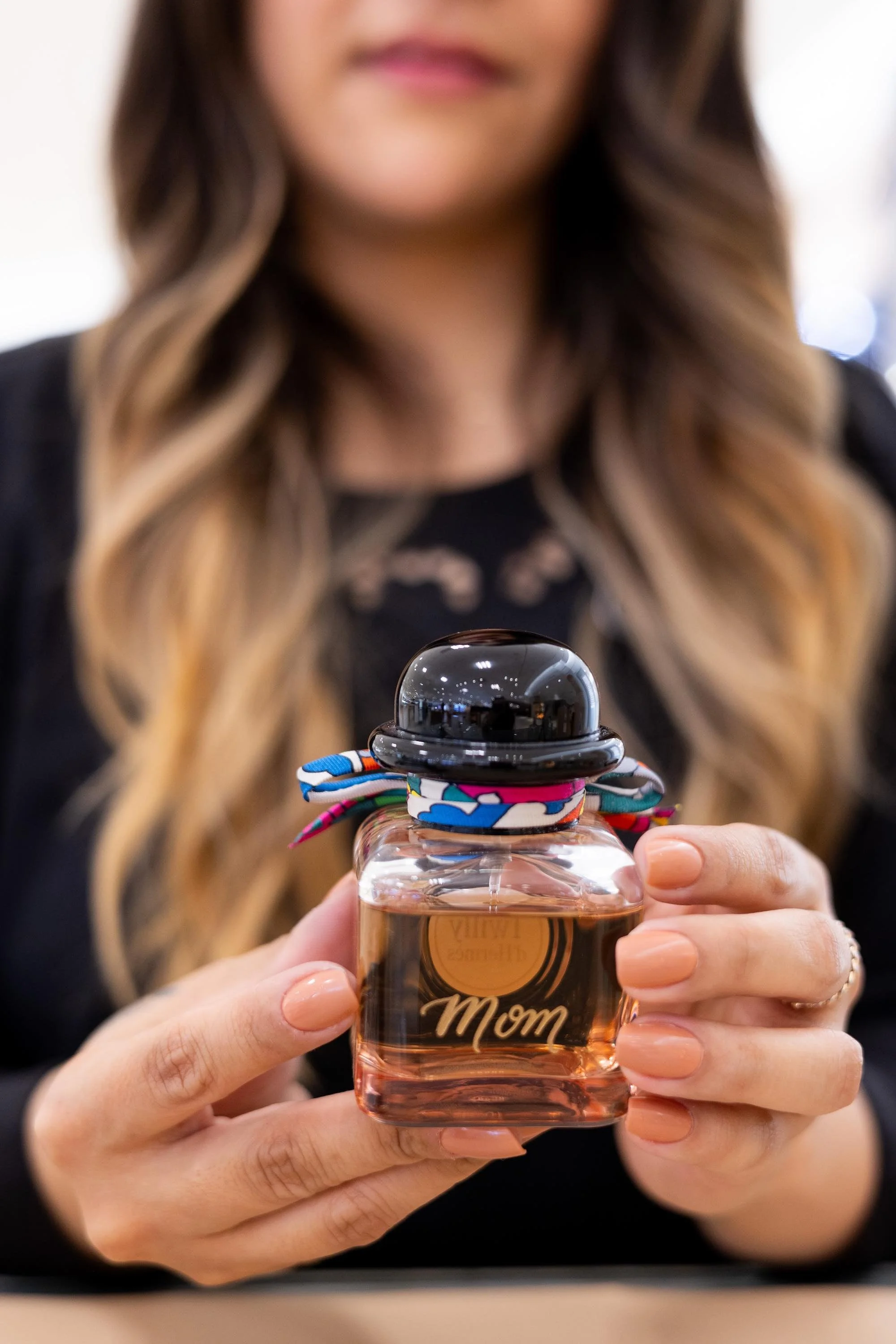 Woman holding a perfume bottle engraved with 'Mom' with a black cap and colorful ribbon tied around neck.
