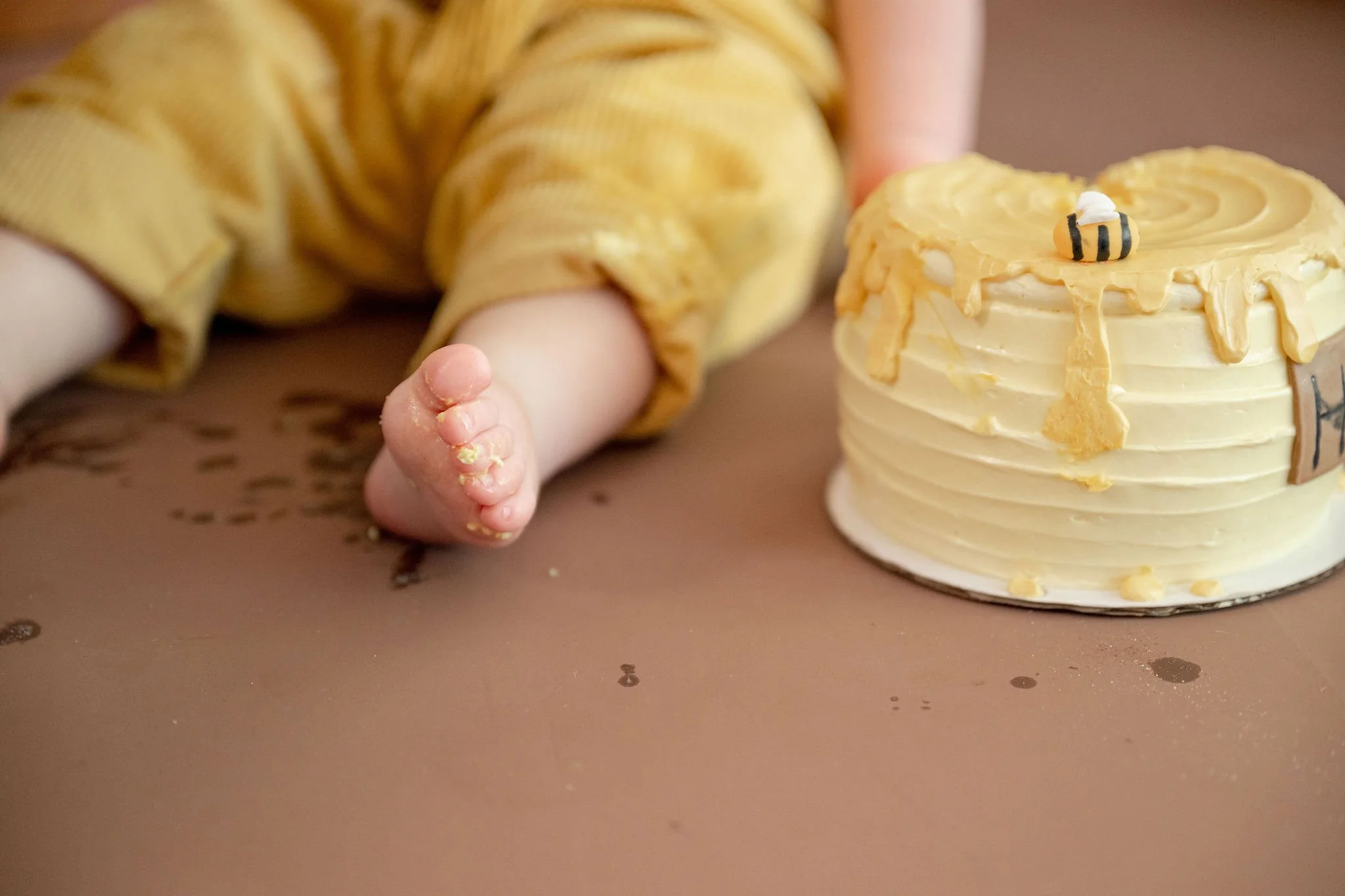 A small child's hand with some finger food, resting next to a yellow birthday cake with a tiny bee decoration on top.