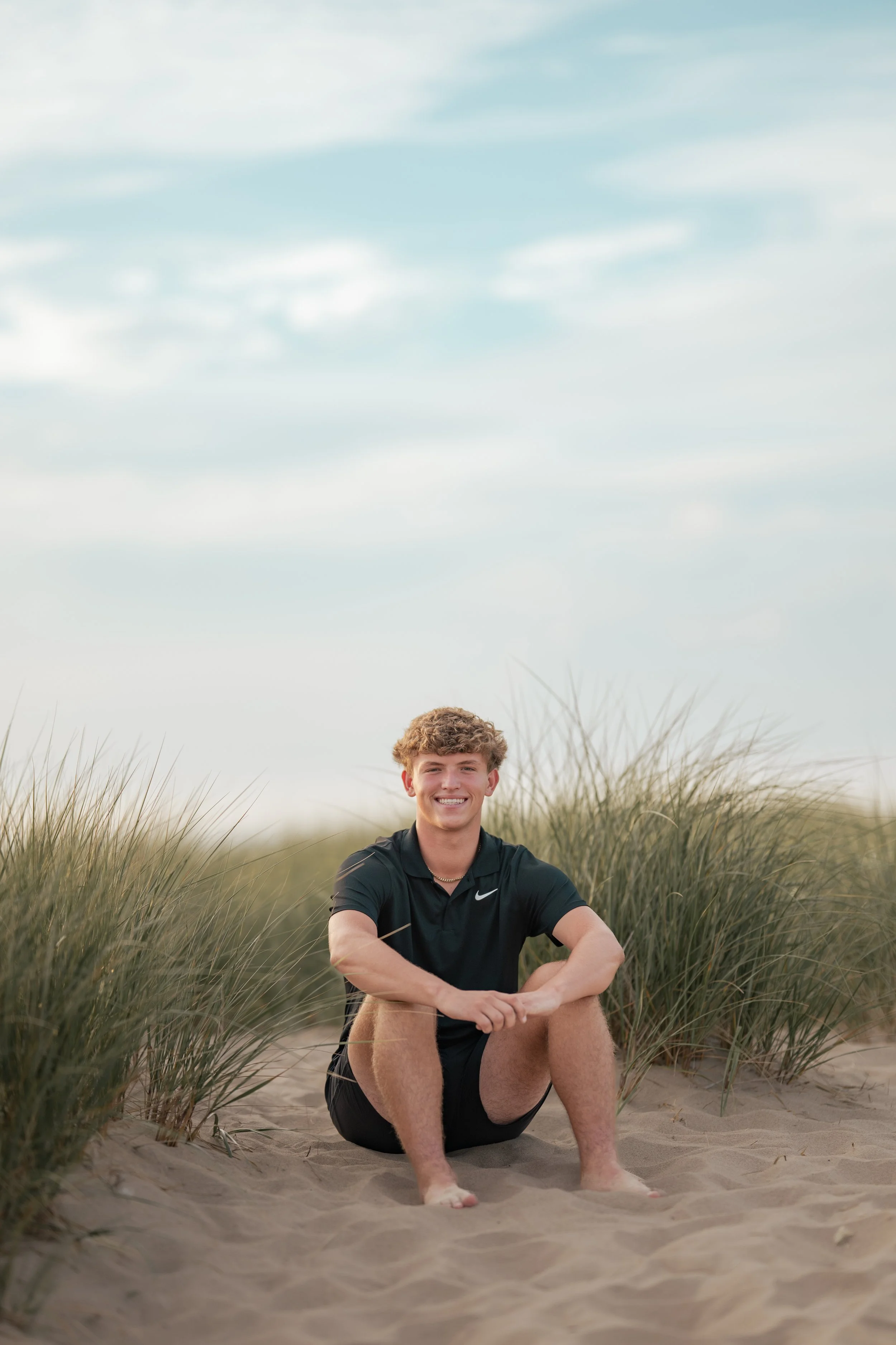 A young man with curly hair and a big smile sitting on sand among tall beach grass, wearing a black Nike shirt, with cloudy sky in the background.