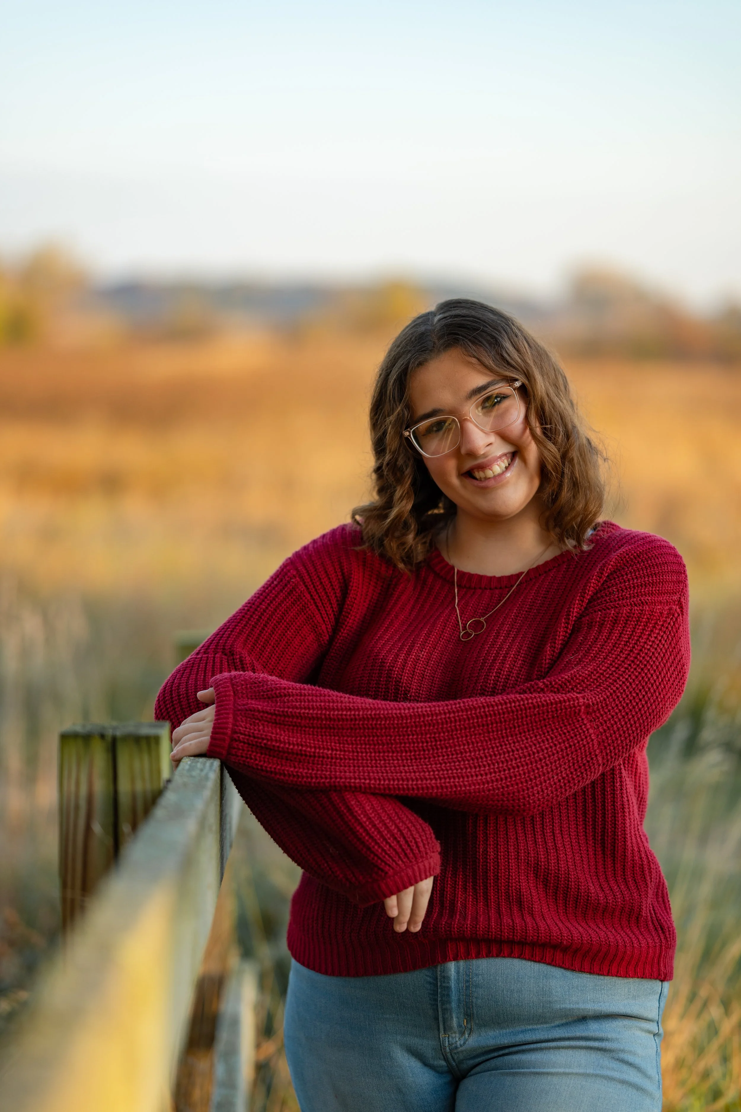 A young woman with shoulder-length curly brown hair and wearing glasses, a red sweater, and blue jeans, smiling outdoors during the daytime with a blurred fall landscape of orange and yellow trees in the background.