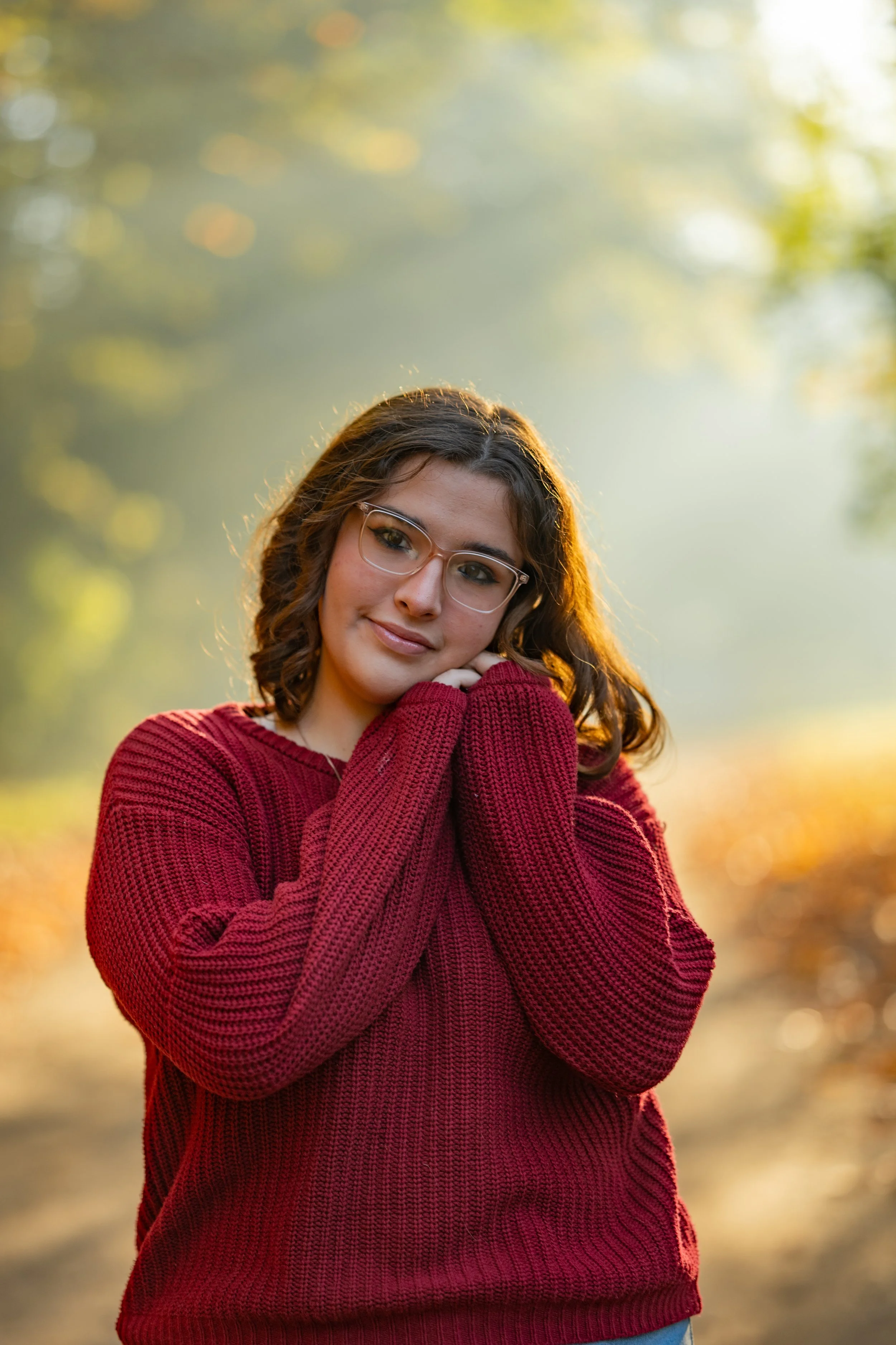 A young woman with curly brown hair, glasses, and a gentle smile, wearing a red knitted sweater, standing outdoors during what appears to be late afternoon or early evening, with a blurred background of trees and sunlight.
