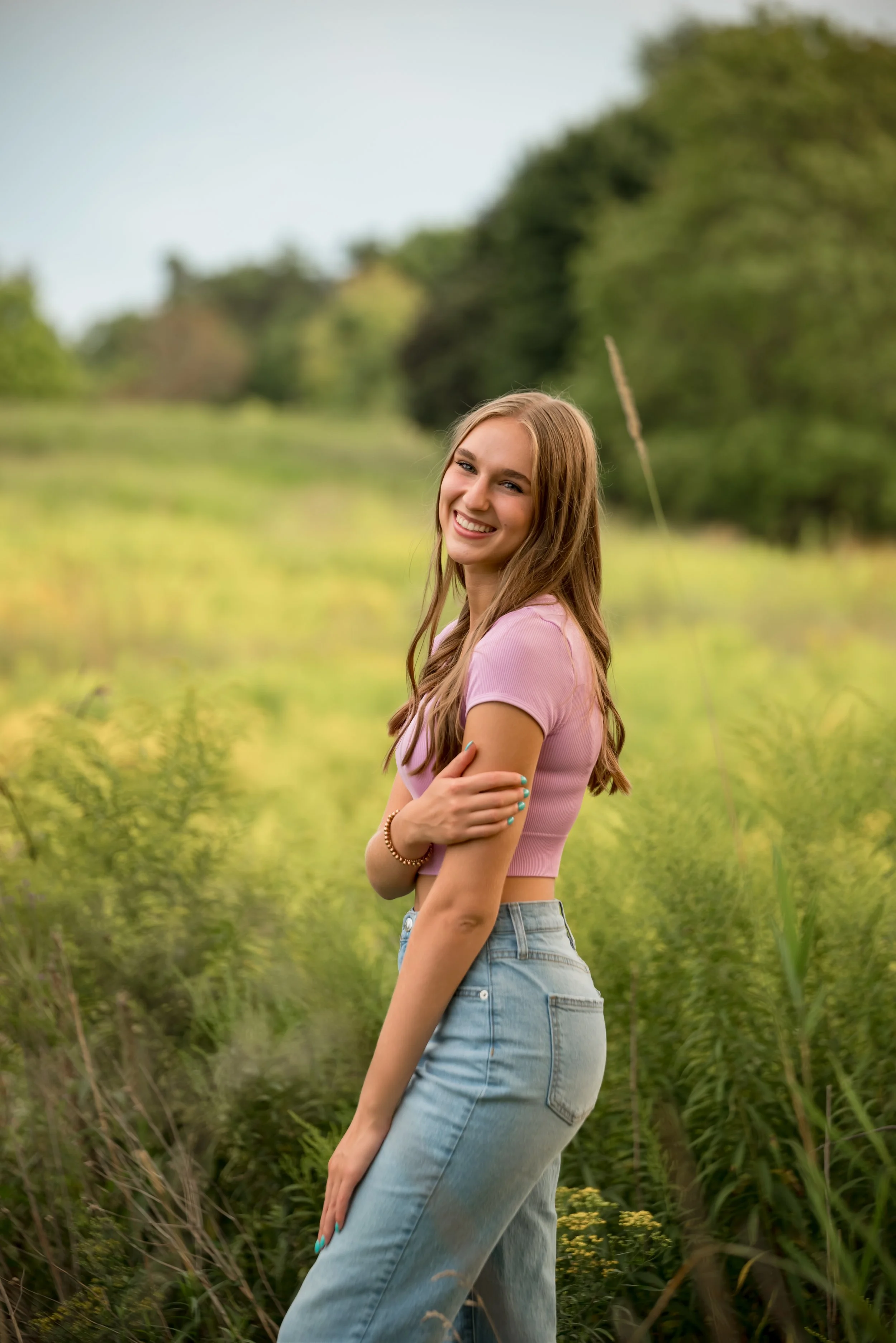 A young woman with long hair smiling and standing in a green, lush outdoor setting, wearing a pink crop top and light blue jeans.
