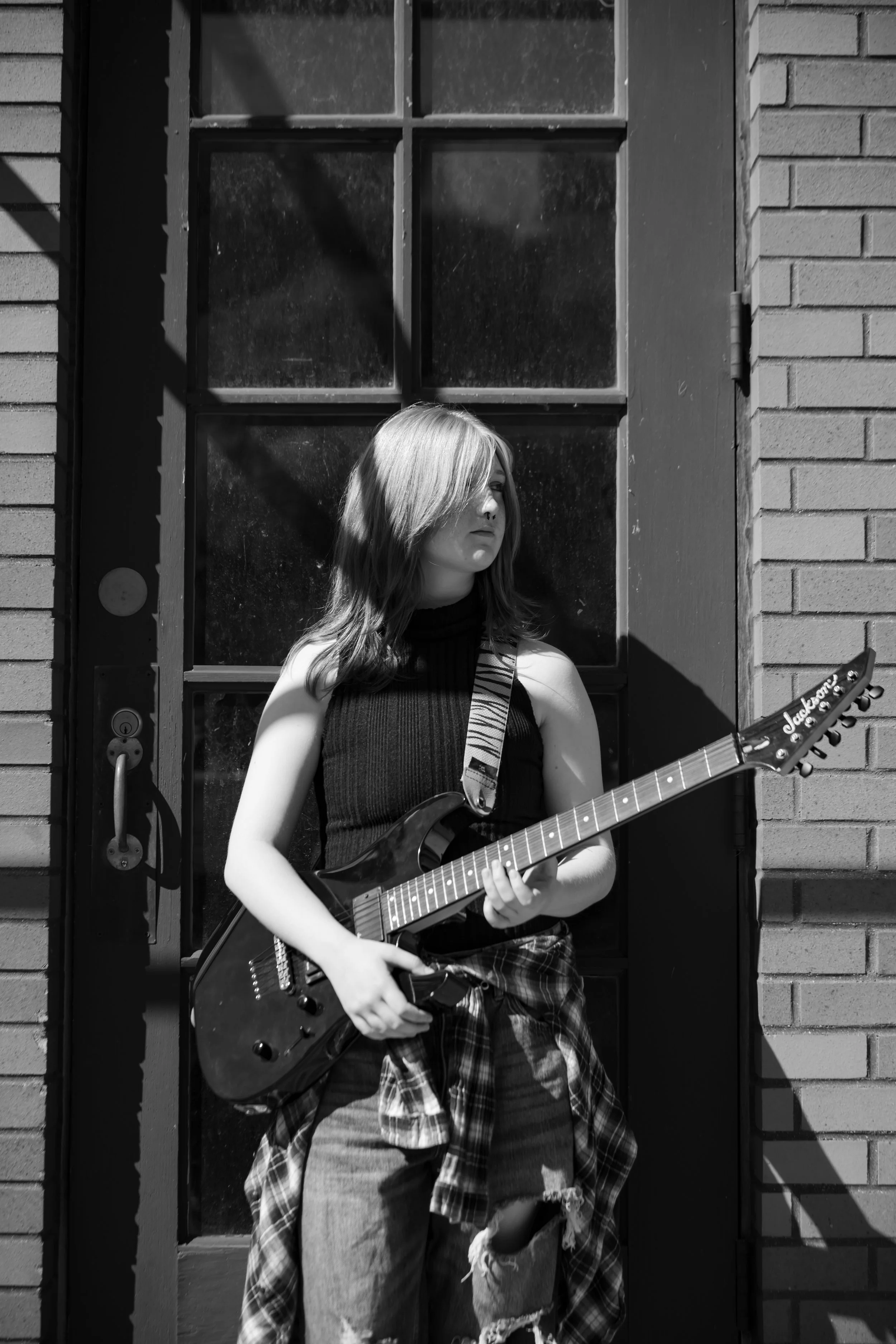 A young woman with long hair playing an electric guitar in front of a brick wall and a window, with sunlight and shadows on her and the wall.