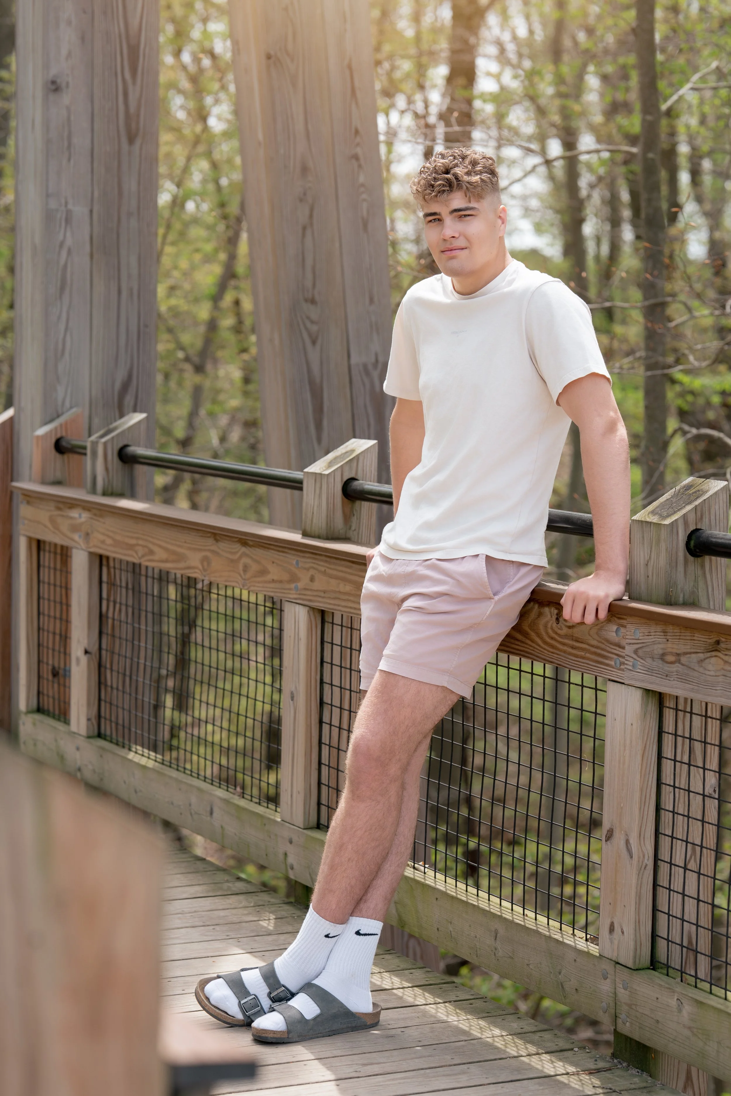 A young man with curly hair leaning on a wooden railing on an outdoor wooden deck in a wooded area, wearing a white T-shirt, light shorts, white socks, and sandals.